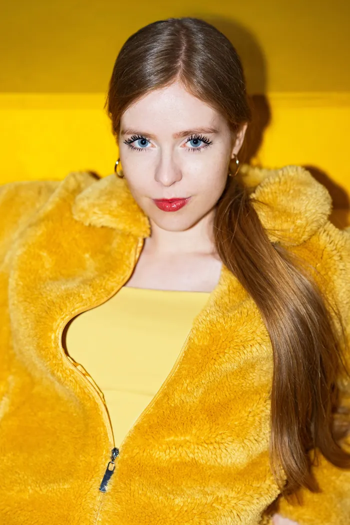 Josefin Schäferhoff in gelber Teddyjacke vor gelbem Hintergrund, roter Lippenstift, selbstbewusster Blick